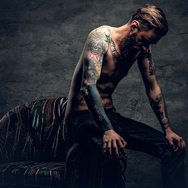 The shirtless, tattooed bearded male sits on a chair over grey background.